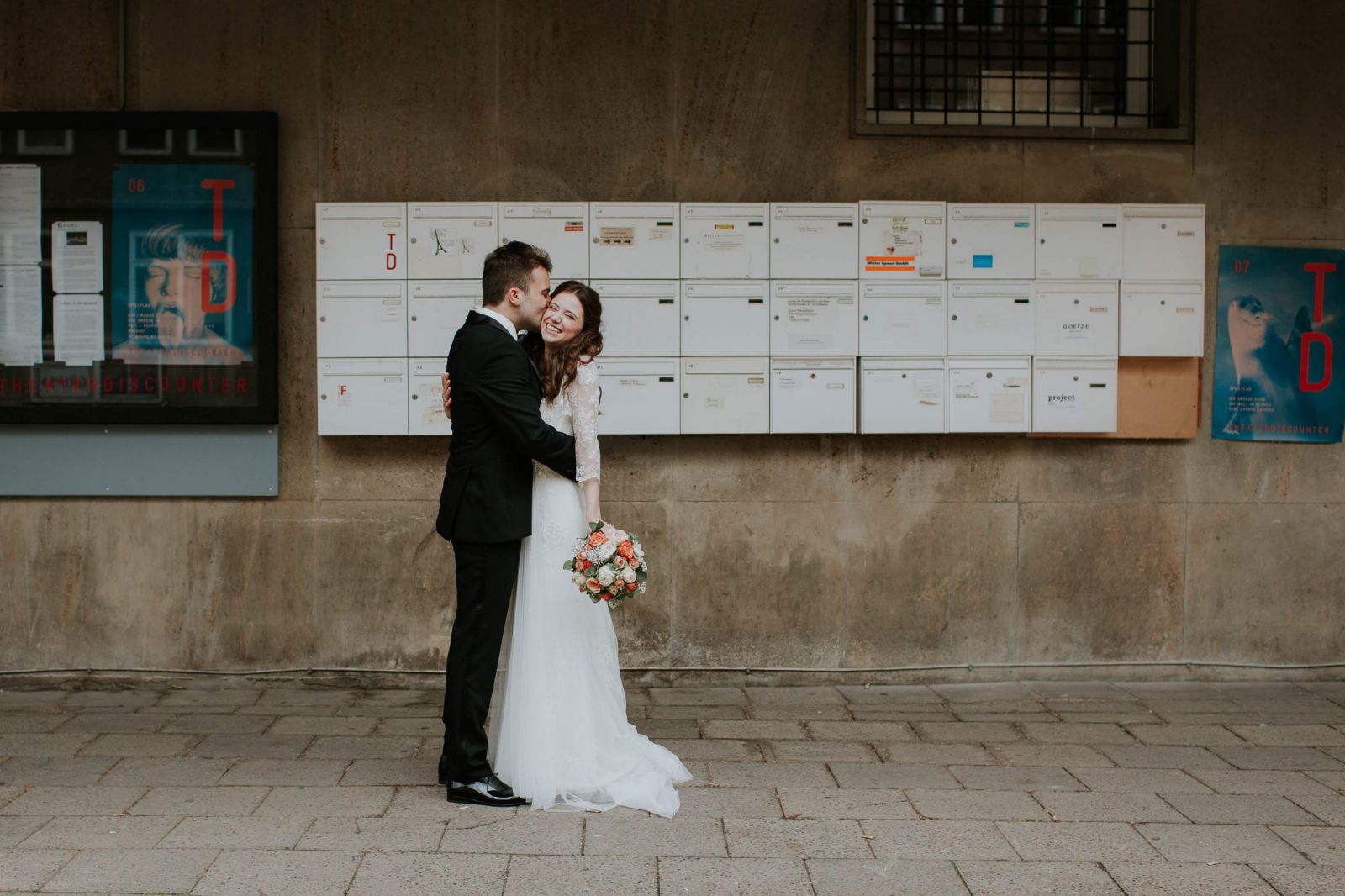 Ein Brautpaar hält sich im Arm, im Hintergrund Briefkästen an einer Hauswand.